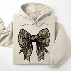 May include: A beige hoodie with a large camouflage bow on the front. The bow is brown and black with a white background.