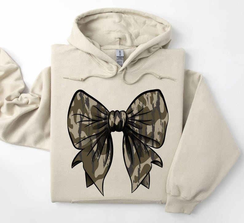 May include: A beige hoodie with a large camouflage bow on the front. The bow is brown and black with a white background.