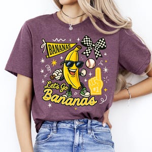 May include: A maroon t-shirt with a graphic of a smiling banana wearing sunglasses, a baseball, and a checkered bow. The text reads "Bananas" and "Let's Go Bananas" in yellow and black.