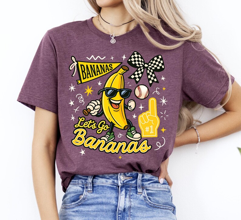 May include: A maroon t-shirt with a graphic of a smiling banana wearing sunglasses, a baseball, and a checkered bow. The text reads "Bananas" and "Let's Go Bananas" in yellow and black.