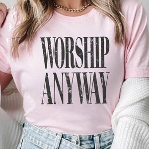 May include: A light pink t-shirt with the words "WORSHIP ANYWAY" printed in large, distressed black letters. The shirt is short-sleeved and made of a soft material.