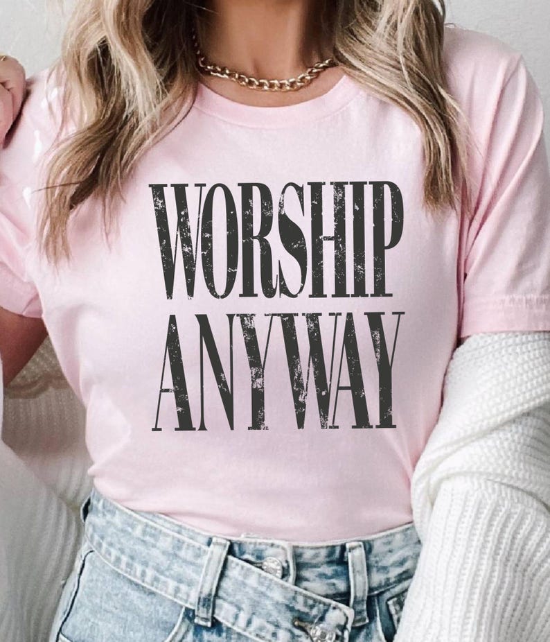 May include: A light pink t-shirt with the words "WORSHIP ANYWAY" printed in large, distressed black letters. The shirt is short-sleeved and made of a soft material.