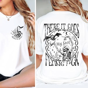 K&ouml;nnte beinhalten: Wei&szlig;es T-Shirt mit einem schwarzen grafischen Aufdruck eines Skeletts, das eine Fledermaus h&auml;lt, und dem Text "There it goes my last flying f*ck".