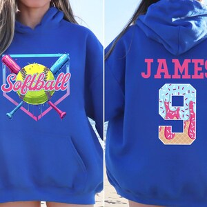 May include: A royal blue hoodie with a softball graphic on the front and the name "JAMES" and the number "9" on the back. The number is decorated with a blue and pink ice cream design.