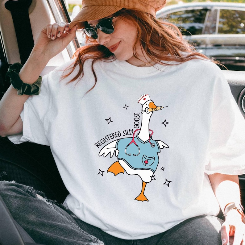K&ouml;nnte beinhalten: Wei&szlig;es T-Shirt mit einer Cartoon-Gans, die eine Krankenschwesterm&uuml;tze und einen Kittel tr&auml;gt. Die Gans h&auml;lt eine Spritze und der Text "Registered Silly Goose" ist auf dem Shirt aufgedruckt.