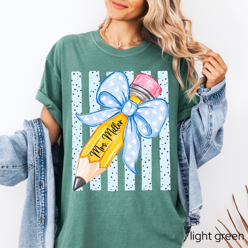 May include: A dark green t-shirt featuring a graphic of a yellow pencil with a pink eraser and a large light blue bow with white hearts. The pencil is printed with the text Mrs. Miller, set against a background of vertical light blue stripes.