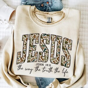 May include: A beige sweatshirt with the word "JESUS" in large, camouflage-patterned letters. Below, the text reads "JOHN 14:6" and "the way the truth the life" in a script font. A gold chain necklace rests on the sweatshirt.