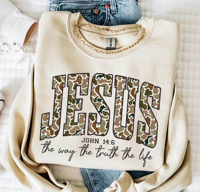 May include: A beige sweatshirt with the word "JESUS" in large, camouflage-patterned letters. Below, the text reads "JOHN 14:6" and "the way the truth the life" in a script font. A gold chain necklace rests on the sweatshirt.