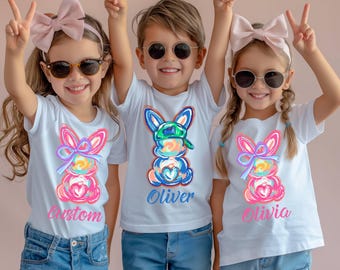 Könnte beinhalten: Drei weiße T-Shirts mit bunten Osterhasendesigns und personalisierten Namen. Die Shirts zeigen einen rosa, blauen und gelben Hasen mit Schleife und grünem Hut. Die Namen "Custom", "Oliver" und "Olivia" sind auf den Shirts aufgedruckt.