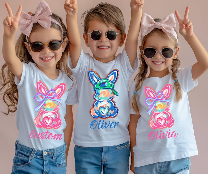 May include: Three white t-shirts with colorful Easter bunny designs and personalized names. The shirts feature a pink, blue, and yellow bunny with a bow and a green hat. The names "Custom", "Oliver", and "Olivia" are printed on the shirts.