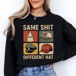 May include: Black sweatshirt with the text "SAME SHIT DIFFERENT HAT" and four images of hats: a KKK hood, a Confederate flag hat, a military hat, and a "Make America Great Again" hat. The design is in a vintage style.