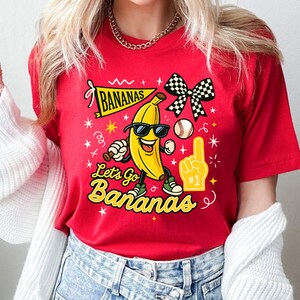 May include: Red t-shirt with a cartoon banana wearing sunglasses and sneakers. The shirt features the text "Bananas" on a banner and "Let's Go Bananas" in yellow, along with a baseball and a foam finger.