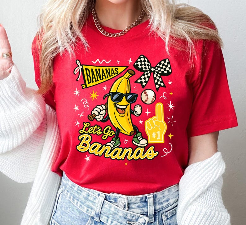 May include: Red t-shirt with a cartoon banana wearing sunglasses and sneakers. The shirt features the text "Bananas" on a banner and "Let's Go Bananas" in yellow, along with a baseball and a foam finger.