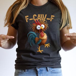 May include: Dark gray t-shirt featuring a cartoon rooster with wide eyes and an open beak. The rooster has a red comb, blue tail feathers, and yellow feet. The text "F-CAW-F" is above the rooster in a yellow font.