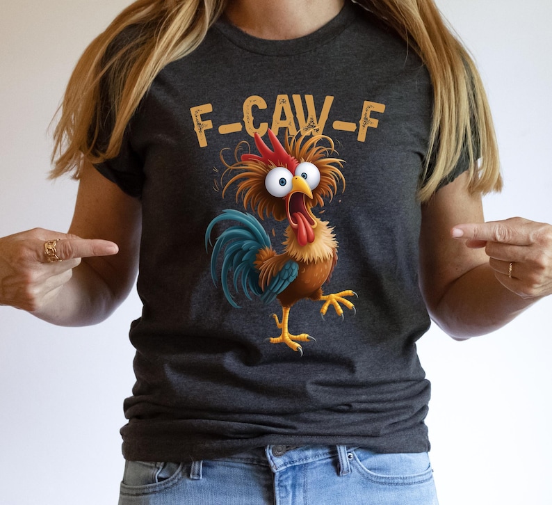 May include: Dark gray t-shirt featuring a cartoon rooster with wide eyes and an open beak. The rooster has a red comb, blue tail feathers, and yellow feet. The text "F-CAW-F" is above the rooster in a yellow font.