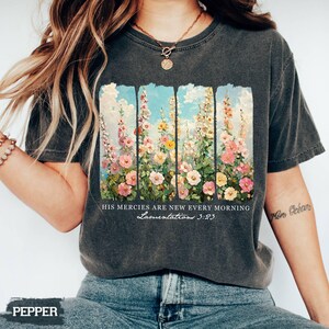 May include: Dark gray t-shirt featuring a floral design with pink, yellow, and white flowers against a blue sky background. The text "HIS mercies are new every morning Lamentations 3:23" is printed below the floral design.