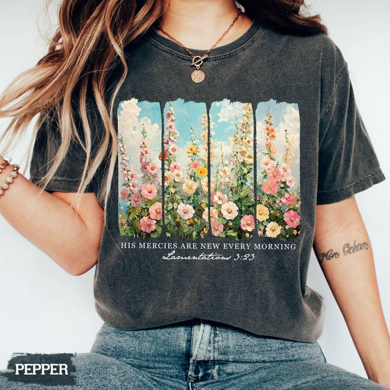 May include: Dark gray t-shirt featuring a floral design with pink, yellow, and white flowers against a blue sky background. The text "HIS mercies are new every morning Lamentations 3:23" is printed below the floral design.