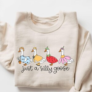 May include: A beige sweatshirt featuring a whimsical illustration of four geese dressed in various outfits, including a straw hat, a yellow cape, and a strawberry costume. The phrase "just a silly goose" is written in cursive below the geese.