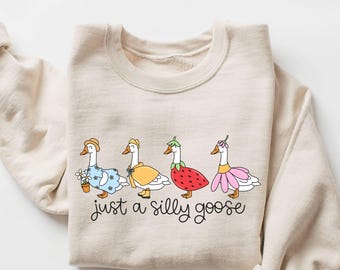 May include: A beige sweatshirt featuring a whimsical illustration of four geese dressed in various outfits, including a straw hat, a yellow cape, and a strawberry costume. The phrase "just a silly goose" is written in cursive below the geese.
