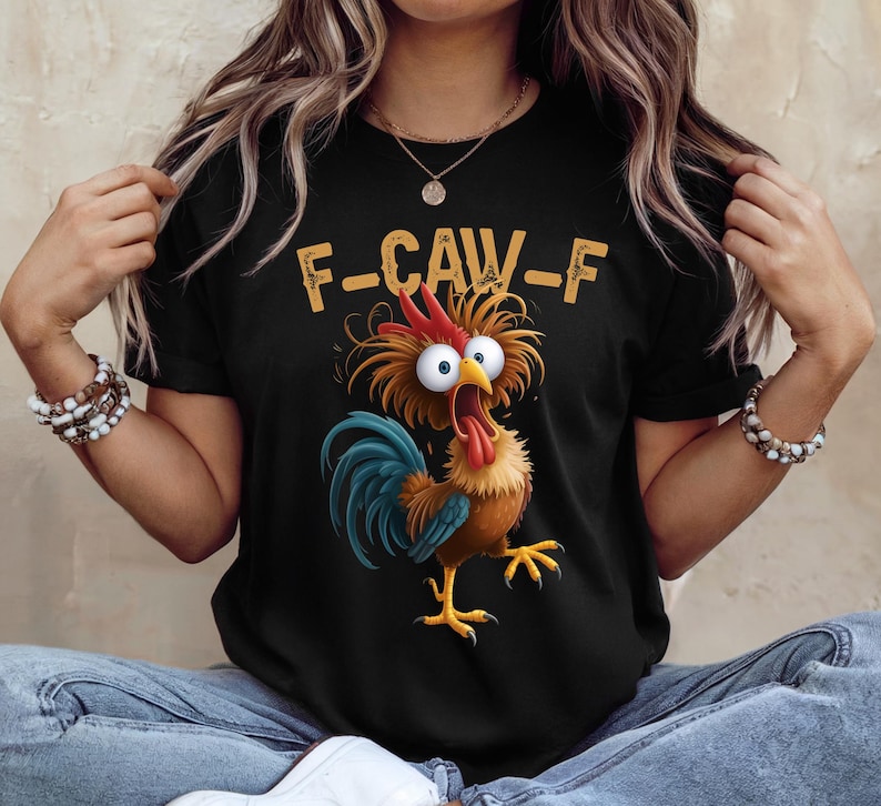 May include: Black t-shirt featuring a cartoon rooster with wide eyes and a long red tongue. The rooster has blue and brown feathers and is positioned below the text "F-CAW-F" in a yellow font. The shirt is a casual wear item.