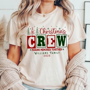 May include: A cream-coloured t-shirt with the words "Christmas Crew" in red and green, along with the text "Making Memories Together" and "Williams Family 2025". The shirt features Christmas tree graphics.