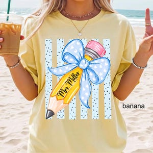 May include: A pale yellow t-shirt featuring a graphic of a yellow pencil with a pink eraser and a large light blue bow with white hearts. The pencil is centered over vertical blue stripes and black dots, with the text Mrs. Miller in black script.