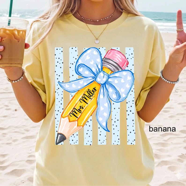 May include: A pale yellow t-shirt featuring a graphic of a yellow pencil with a pink eraser and a large light blue bow with white hearts. The pencil is centered over vertical blue stripes and black dots, with the text Mrs. Miller in black script.
