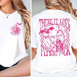 K&ouml;nnte beinhalten: Wei&szlig;es T-Shirt mit einem rosafarbenen Grafikdruck auf Vorder- und R&uuml;ckseite. Die Vorderseite zeigt den Aufdruck "there it goes" mit einer kleinen Fledermaus und Blumen. Die R&uuml;ckseite zeigt den Aufdruck "There it goes my last flying f*ck" mit einem Skelett, das eine Fledermaus und Blumen h&auml;lt.