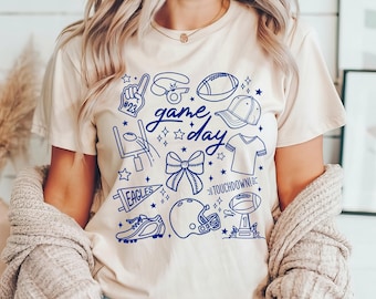 May include: A cream-coloured t-shirt with a blue graphic design featuring football-related illustrations and the words "game day." The design includes a football, helmet, and other sports-themed elements. The shirt is made of a soft material.