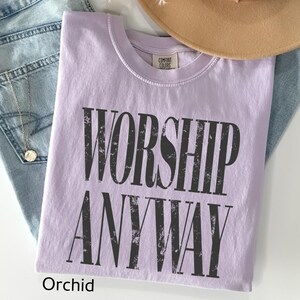 May include: Orchid-colored t-shirt with the words "WORSHIP ANYWAY" in distressed black lettering. The shirt is folded and rests on a pair of blue jeans. A tan hat is visible in the background.