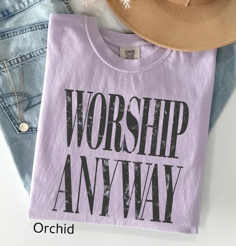 May include: Orchid-colored t-shirt with the words "WORSHIP ANYWAY" in distressed black lettering. The shirt is folded and rests on a pair of blue jeans. A tan hat is visible in the background.