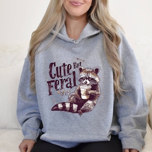 May include: Gray hooded sweatshirt with a raccoon graphic holding a coffee cup and the text "Cute But Feral".