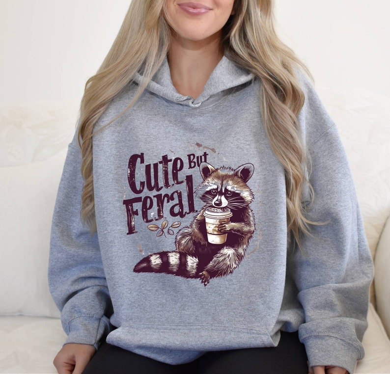 May include: Gray hooded sweatshirt with a raccoon graphic holding a coffee cup and the text "Cute But Feral".