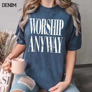 May include: Denim blue t-shirt with the words "WORSHIP ANYWAY" in large white block letters. The shirt is worn by a person holding a pink mug. The word "DENIM" is printed in black in the upper left corner.