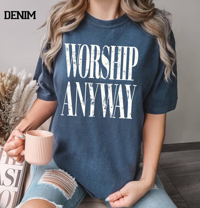 May include: Denim blue t-shirt with the words "WORSHIP ANYWAY" in large white block letters. The shirt is worn by a person holding a pink mug. The word "DENIM" is printed in black in the upper left corner.