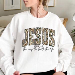 May include: White sweatshirt with the word "JESUS" in a camouflage pattern across the chest. Below the word "JESUS" is the text "JOHN 14:6" and "the way the truth the life" in black font.