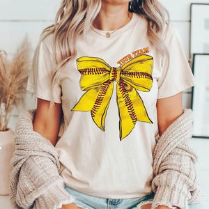 May include: A cream-coloured t-shirt with a yellow and red softball bow graphic and the text "Your Team".