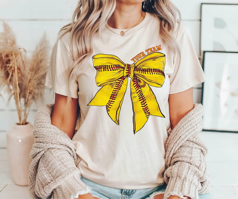 May include: A cream-coloured t-shirt with a yellow and red softball bow graphic and the text "Your Team".