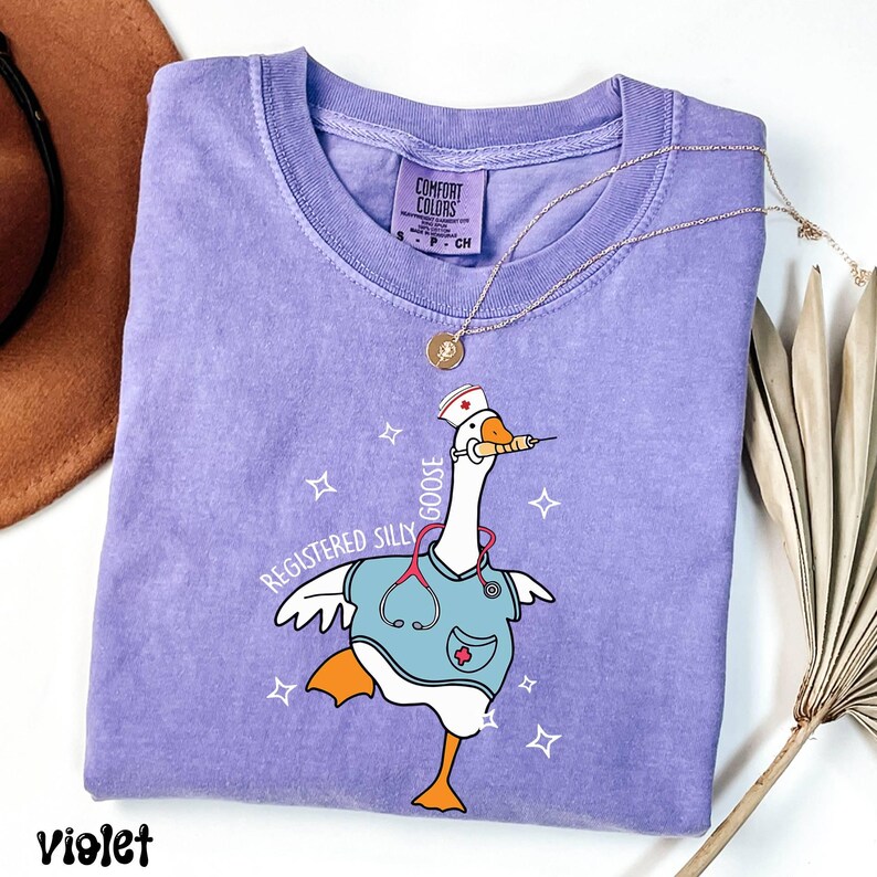 K&ouml;nnte beinhalten: Lilafarbenes T-Shirt mit einer Cartoon-Gans, die eine Krankenschwesterm&uuml;tze und einen Kittel tr&auml;gt. Die Gans h&auml;lt eine Spritze und der Text "Registered Silly Goose" ist auf dem T-Shirt aufgedruckt.
