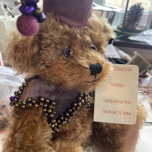 May include: A brown mohair teddy bear wearing a purple hat with a purple pom-pom and a purple and gold beaded necklace. The bear is handmade by Emma and is called Periwinkle.