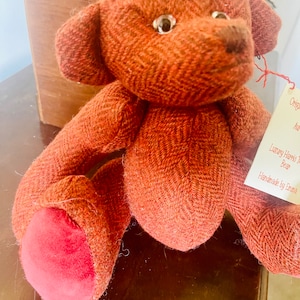 May include: A handmade brown tweed teddy bear with red felt feet. The bear is sitting on a wooden surface. The tag reads "Luxury Harris Tweed Bean Handmade by Emma".