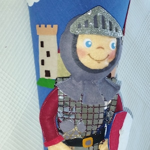 Könnte beinhalten: Ein blauer Filz-Kegel-Beutel mit dem Namen "JOHANN" in weißen Buchstaben. Er zeigt einen Ritter aus Filz mit silbernem Helm, der ein Schwert und einen Schild hält. Ein Schloss und Wolken sind ebenfalls abgebildet.