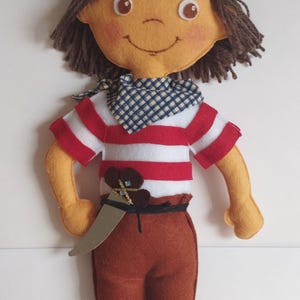 Könnte beinhalten: Eine handgefertigte Filzpuppe, die als Pirat verkleidet ist. Die Puppe hat braunes Haar, ein rot-weiß gestreiftes Hemd, eine braune Hose und einen schwarzen Piratenhut mit einem weißen Totenkopf und Knochen.
