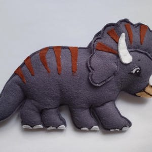 Könnte beinhalten: Ein dunkelgraues Triceratops-Dinosaurier-Stofftier aus Filz mit rostfarbenen Streifen auf dem Rücken, weißen Hörnern und weißen Krallen. Der Dinosaurier hat einen großen Nackenschild, eine hellbraune Schnauze und ein schwarzes Auge mit weißem Glanzpunkt.