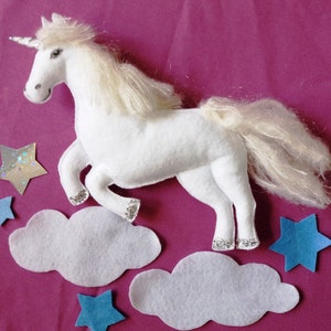 Puede incluir: Un unicornio blanco de fieltro con cuerno, melena y pezuñas plateadas. El unicornio está rodeado de nubes de fieltro blanco, estrellas de fieltro azul y una estrella de purpurina plateada sobre un fondo rosa. Una artesanía caprichosa y hecha a mano.