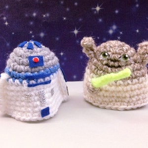 Könnte beinhalten: Zwei gehäkelte Star Wars-Charaktere, R2-D2 und Yoda, stehen auf einer weißen Oberfläche. R2-D2 ist blau, weiß und grau, während Yoda beige und grün ist. Yoda hält ein grünes Lichtschwert.