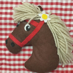 Puede incluir: Una cabeza de caballo de fieltro marrón con una brida roja y una melena de hilo color crema. Una margarita blanca y amarilla decora la cabeza del caballo. La cabeza de caballo está sobre un fondo a cuadros rojos y blancos.