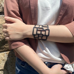 Puede incluir: Un brazalete geométrico negro con un diseño de corte. El brazalete se lleva en la muñeca de una persona que lleva una chaqueta marrón y una camisa blanca.