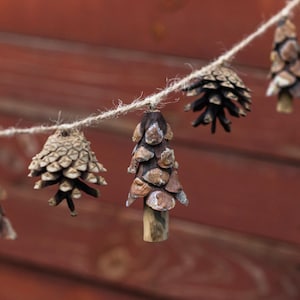 Puede incluir: Una guirnalda rústica con piñas y adornos de piñas ensartados en una cuerda natural. Las piñas son de varios tamaños y formas, con tonos marrones y beige naturales. El fondo es una superficie de madera marrón rojizo.