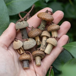Puede incluir: Una colección de pequeños colgantes con forma de seta hechos de materiales naturales. Cada pieza presenta una tapa de bellota texturizada y un tallo de madera, unidos a un cordón marrón fino. Los colgantes varían en tonos marrones y beige.